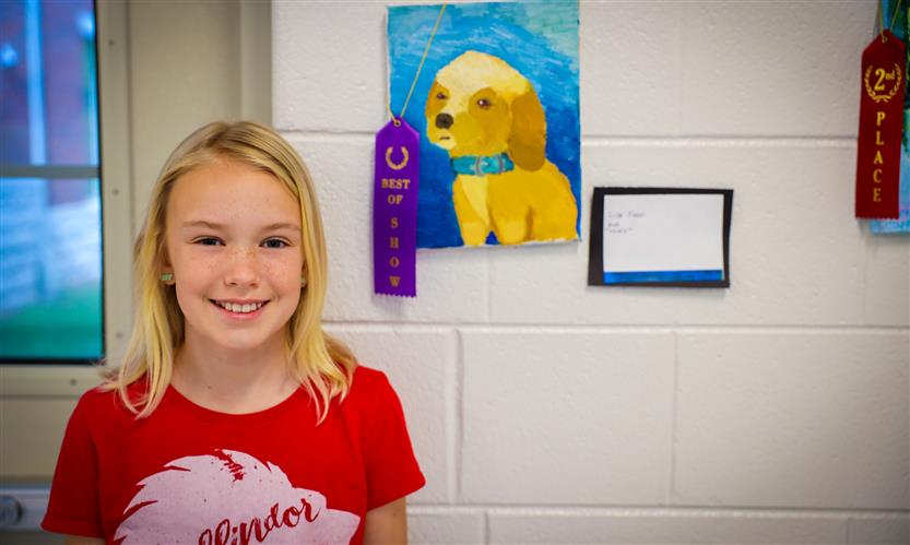 A girl wearing a red shirt smiles in front of a drawing of a dog